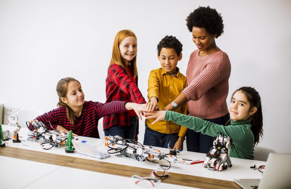 Group of happy kids with their African American female science teacher programming electric toys and robots at robotics classroom